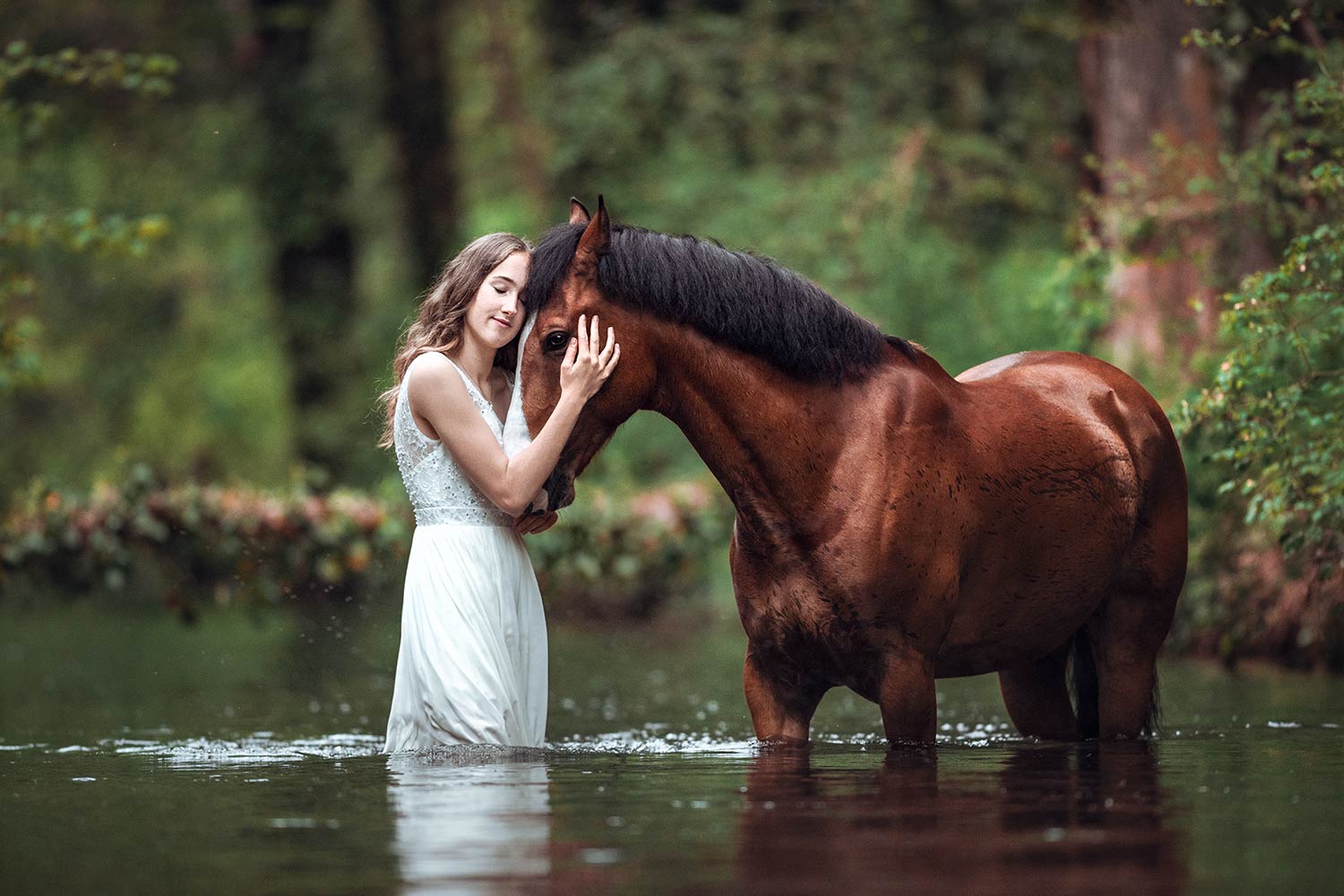 Eine junge Frau in einem weissen Kleid umarmt liebevoll ein dunkelbraunes Pferd, das im Wasser steht.