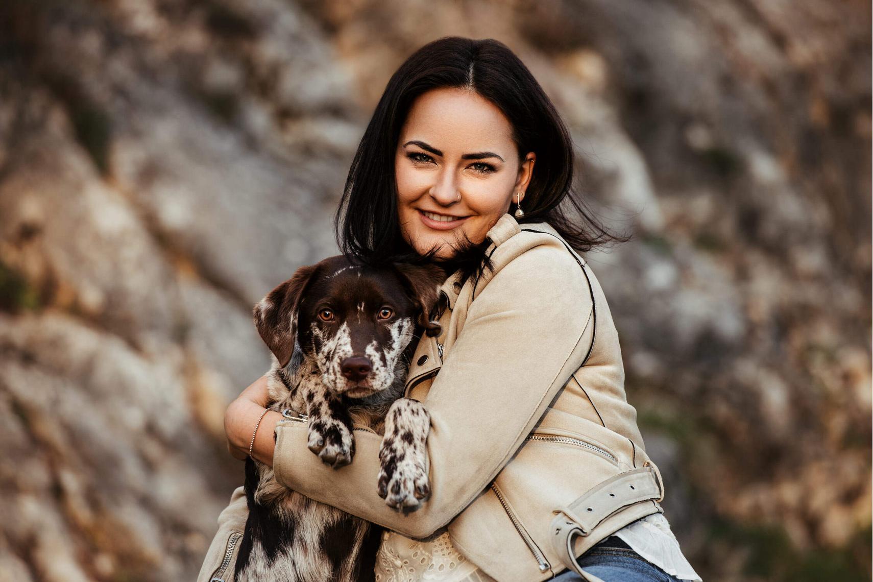 Eine junge Frau umarmt ihren gesprenkelten Hund liebevoll, während sie vor einem steinigen Hintergrund posieren.