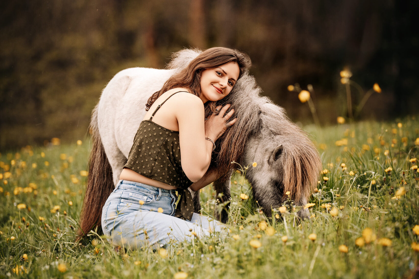 Lächelnde Frau schmiegt sich in einer Löwenzahnwiese an ein kleines graues Sherland Pony